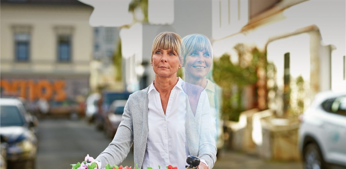 Eine Frau mit einem Fahrrad und einem bunten Blumenstrauß fährt durch eine Straße, während im Hintergrund eine unscharfe Darstellung zu sehen ist.
